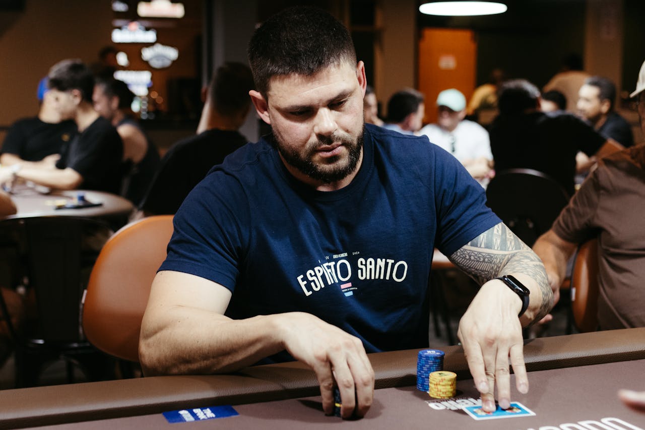 An adult man plays poker at a casino table, focusing on his hand and poker chips.